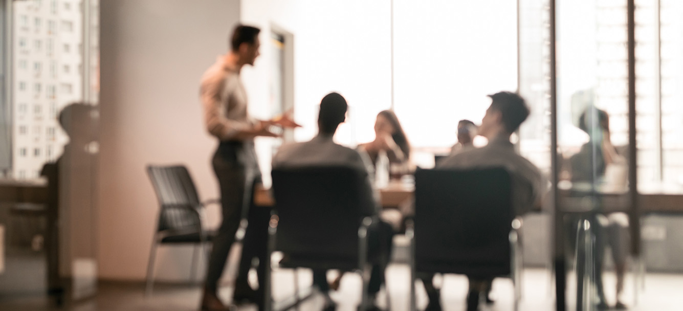 Business Presentation, Blurred Background. Businessman Giving Speech During Seminar With Coworkers In Office, Standing At Desk In Boardroom, Diverse People Sitting At Table And Listening To Speaker